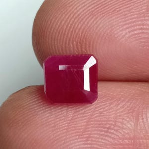 May include: A faceted, rectangular ruby gemstone with a deep red hue. The gem is held between fingers, showcasing its polished surfaces and internal clarity. The ruby's shape is a modified emerald cut, with a slightly rounded rectangular form.