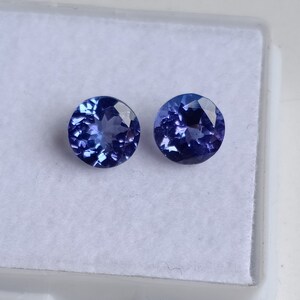 May include: Two round, faceted, blue gemstones. The gemstones are set in a white, velvet-lined box.