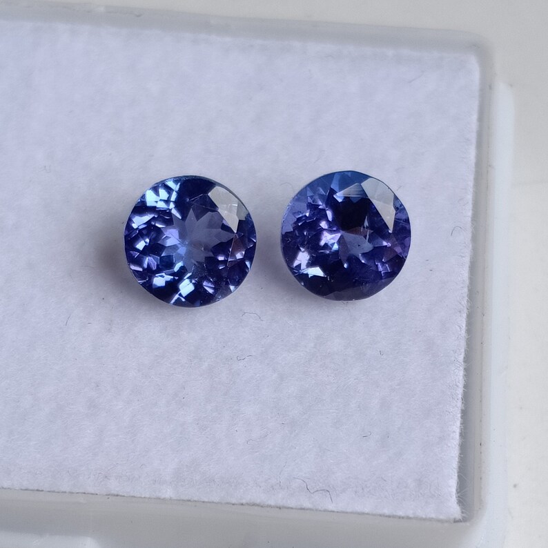 May include: Two round, faceted, blue gemstones. The gemstones are set in a white, velvet-lined box.