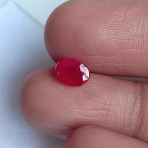 May include: A faceted, oval-shaped, deep red gemstone. The gem is held between two fingers, showcasing its polished surface and internal reflections. The stone's color is a vibrant, saturated red, typical of a ruby.