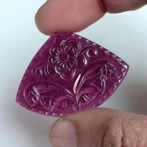 May include: A magenta-coloured, triangular gemstone with a carved floral pattern. The design includes a central flower and surrounding leaves. The gemstone is held between two fingers.