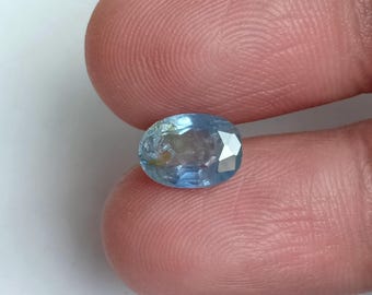 Könnte beinhalten: Ein ovalförmiger, hellblauer Edelstein mit Facetten. Der Edelstein hat ein leicht trübes Aussehen mit einigen inneren Einschlüssen. Der Stein wird zwischen zwei Fingern gehalten, was seine Größe und Farbe zeigt. Wahrscheinlich ein Saphir oder Aquamarin.