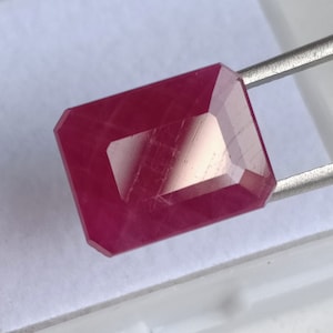 Pode incluir: Uma gema vermelha escura com um corte esmeralda retangular. A gema é segurada por uma pinça.