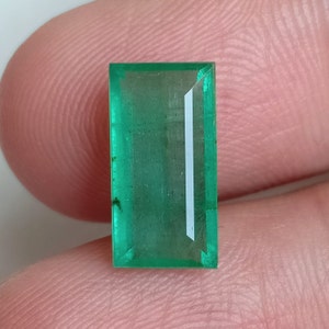 Op de afbeelding: Een levendige groene smaragd edelsteen met een rechthoekige snede en een kleine witte insluiting.