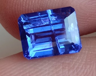 May include: A rectangular, faceted gemstone with a deep blue hue. The gem has a series of parallel cuts across its surface, creating a grid-like pattern. The facets reflect light, giving the gem a brilliant appearance. The gem is held between fingers.