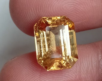 May include: A golden yellow gemstone with a rectangular emerald cut. The gemstone is faceted and has a slight shimmer.