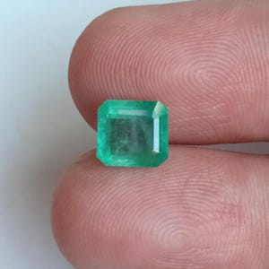 May include: A square-cut emerald gemstone, displaying a vibrant green hue. The gemstone is held between two fingers, showcasing its clarity and facets. The emerald's shape is a classic rectangular cut with trimmed corners.