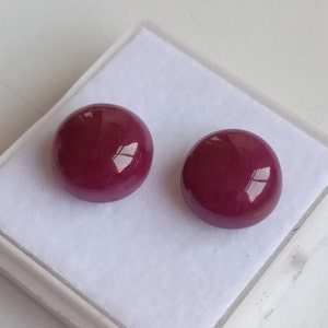 Puede incluir: Dos cabujones redondos y pulidos, de color burdeos, se muestran en una caja blanca. Los cabujones tienen un acabado brillante, que refleja la luz. Estos cabujones se utilizan probablemente para la fabricación de joyas u otros proyectos de artesanía.