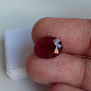 Könnte beinhalten: Ein facettierter, oval geformter Rubin-Edelstein. Der Edelstein hat eine tiefrote Farbe und scheint zwischen Fingern gehalten zu werden. Der Rubin ist vor einem weißen Hintergrund platziert.