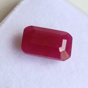 May include: A deep red gemstone with a rectangular emerald cut. The gemstone is faceted and has a high polish.