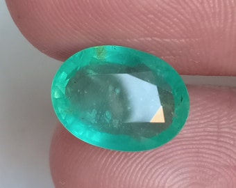 May include: A vibrant green emerald gemstone with a faceted oval cut. The stone has a slight cloudy appearance and is set in a ring.