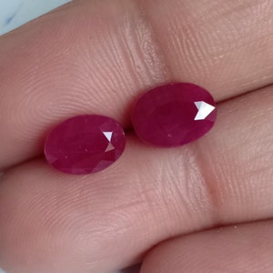 Könnte beinhalten: Zwei ovale Rubin-Edelsteine, jeder mit einer tief roten Farbe und einem facettierten Schliff.