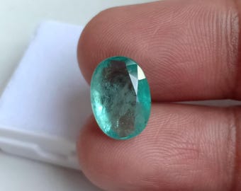May include: An oval-shaped, light teal gemstone with cut facets. The gem is held between fingers, highlighting its polished surface and internal clarity. The stone's colour is a vibrant, cool teal, reflecting light.