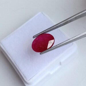 Puede incluir: Una gema de rubí facetada, de forma ovalada, sujeta con pinzas plateadas. El rubí es de color rojo intenso y se encuentra sobre una superficie blanca. La gema es el punto focal, mostrando sus facetas pulidas y su vibrante tonalidad.