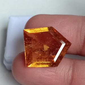 Könnte beinhalten: Ein großer, facettierter, orangefarbener Edelstein mit sechseckiger Form. Der Edelstein hat ein leicht trübes Aussehen und ist in eine klare, facettierte Fassung gefasst.