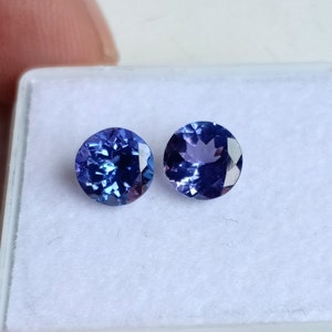 May include: Two round, faceted, blue gemstones with a purple hue. The gemstones are set in a white box.