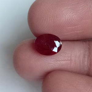 Pode incluir: Uma pedra preciosa de rubi facetada em forma oval. A gema é de uma cor vermelha profunda e rica com uma superfície polida. As facetas captam a luz, criando reflexos brilhantes. O rubi é segurado entre dois dedos.