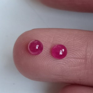 May include: Two round, pink ruby gemstones, each with a smooth, polished surface, are displayed on a finger.