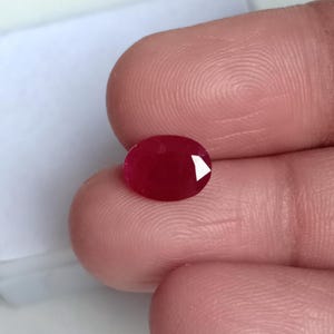 May include: A faceted, oval-shaped ruby gemstone. The gem is a deep red colour with a polished surface and a bright, reflective facet. The ruby is held between two fingers, with a soft, blurred background.