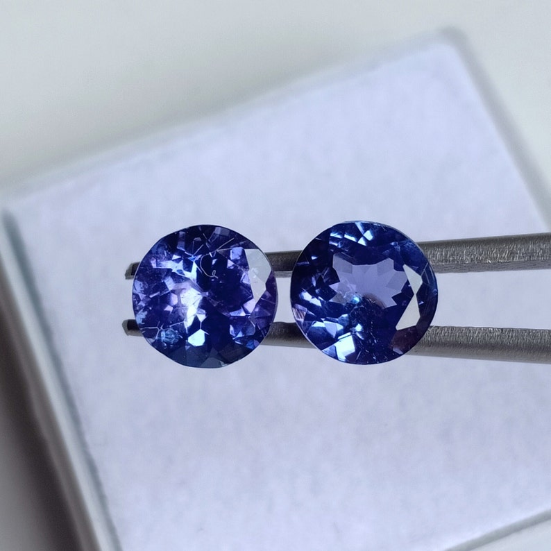 May include: Two round, faceted, blue gemstones with a purple hue. The gemstones are held by a pair of tweezers.