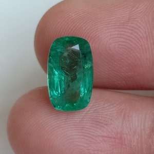 Puede incluir: Una gema de esmeralda verde vibrante con un corte rectangular. La esmeralda tiene una superficie lisa y pulida y está engastada en un engaste claro y facetado.