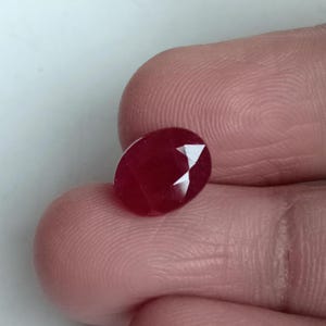 May include: A faceted, oval-shaped ruby gemstone. The ruby is a deep red color with a polished surface and several facets that reflect light. The gemstone is held between two fingers.