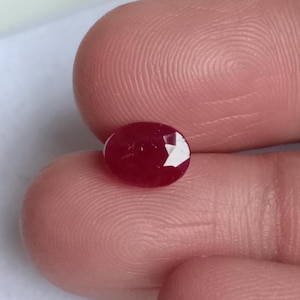 Może przedstawiać: Fasetowany, owalny kamień szlachetny rubinu. Rubin ma głęboki, bogaty czerwony kolor z wypolerowaną powierzchnią i kilkoma widocznymi fasetami. Kamień szlachetny jest trzymany między dwoma palcami.