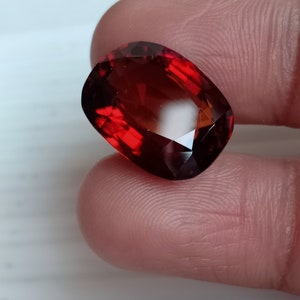 May include: A deep red gemstone with a faceted cut. The gemstone is oval shaped and has a high polish.