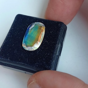 May include: A clear oval-shaped gemstone with iridescent colors, including blue, green, and yellow, displayed in a black velvet box.