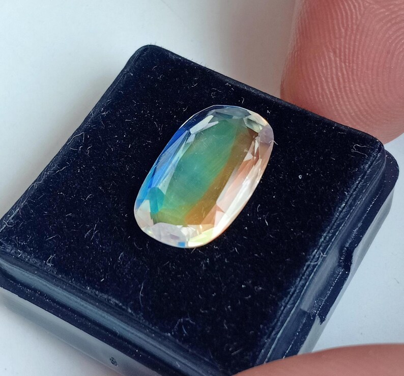 May include: A clear oval-shaped gemstone with iridescent colors, including green, blue, and orange, displayed in a black velvet box.