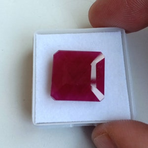 Puede incluir: Una piedra preciosa de color rojo intenso con un corte cuadrado esmeralda, expuesta en una caja blanca.