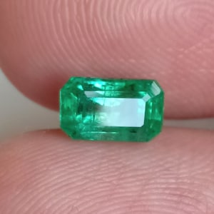 May include: A rectangular emerald gemstone with a vibrant green hue. The gem has a faceted cut, showcasing its clarity and brilliance. The emerald is held between two fingers, highlighting its size and shape.