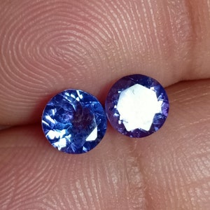 May include: Two round, faceted, blue gemstones with a purple hue. The gemstones are approximately 5mm in diameter.