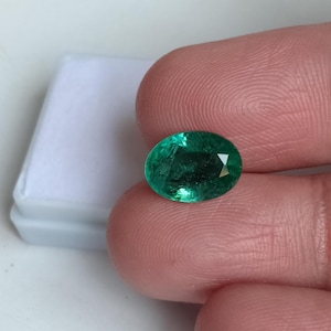 May include: Oval-cut emerald gemstone, a vibrant green hue, held between fingers. The gem is set against a white background, showcasing its clarity and brilliance. A classic gemstone for jewelry.