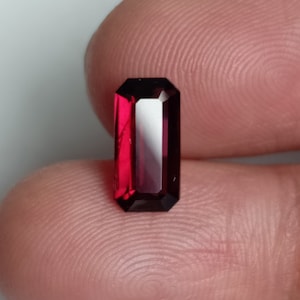 May include: A deep red gemstone with a rectangular emerald cut. The gemstone is faceted and has a high polish.