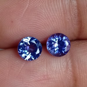 May include: Two round, blue gemstones with a faceted cut. The gemstones are set against a light pink background.