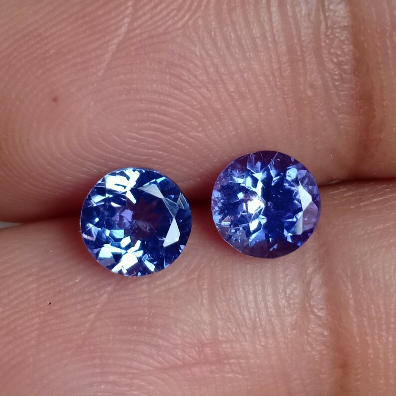 May include: Two round, blue gemstones with a faceted cut. The gemstones are set against a light pink background.