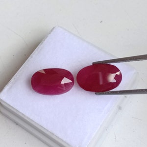 Puede incluir: Dos piedras preciosas ovaladas de color rojo con un corte facetado. Las piedras preciosas están colocadas en una caja blanca y se sostienen con unas pinzas.
