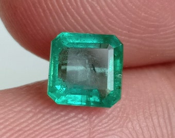 Puede incluir: Una gema de esmeralda verde vibrante, cortada en forma cuadrada con bordes biselados. La piedra es clara y tiene un ligero brillo.