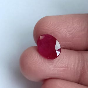 May include: A faceted, oval ruby gemstone. The ruby is a deep red colour with multiple facets that reflect light. The gemstone is held between two fingers.
