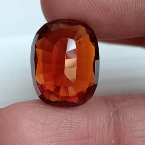 May include: A deep orange oval-shaped gemstone with a faceted cut. The gemstone is set against a light pink background.