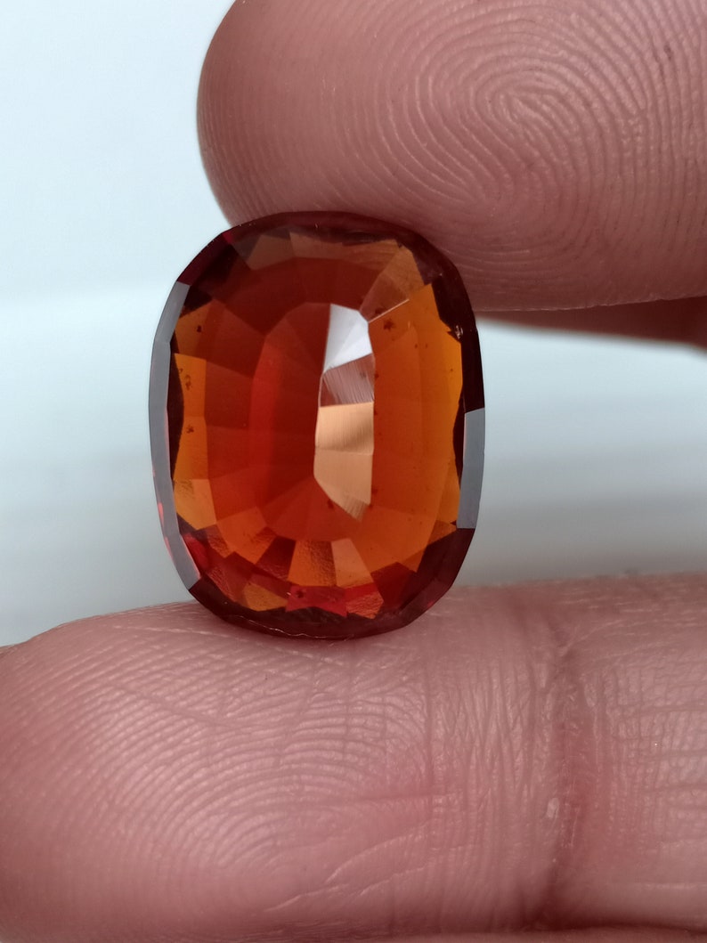 May include: A deep orange oval-shaped gemstone with a faceted cut. The gemstone is set against a light pink background.