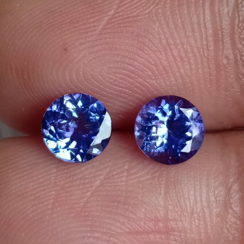 May include: Two round, blue gemstones with a faceted cut. The gemstones are set against a pink background.