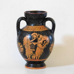 Puede incluir: Un jarrón de cerámica negro con dos asas, con ilustraciones en naranja y negro de figuras griegas antiguas. El jarrón tiene un borde decorativo y una base de pedestal. La obra de arte representa guerreros y otras figuras.