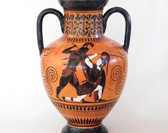 May include: A terracotta-colored ceramic amphora featuring black-figure style illustrations of two warriors in combat. The vessel has two black handles, a flared rim, and a pedestal base, with decorative geometric patterns encircling the neck and base.