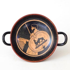 Può includere: Un kylix in ceramica nera e terracotta con un'illustrazione centrale di una figura barbuta che tiene una lancia e uno scudo rotondo. Lo scudo mostra la sagoma di un animale a quattro zampe. La ciotola ha due manici neri e un bordo rosso.