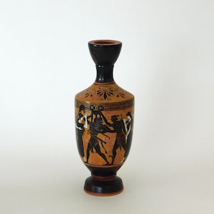 May include: A small, brown ceramic vase with black and red painted figures on the side. The vase has a narrow neck and a wide base. The figures are of people in a scene from Greek mythology.