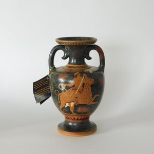 Puede incluir: Una ánfora de cerámica verde oscuro con dos asas y una base de pedestal. El jarrón presenta una escena pintada de una figura montada en un animal manchado, con detalles en rojo y naranja. Una etiqueta está adherida al jarrón.