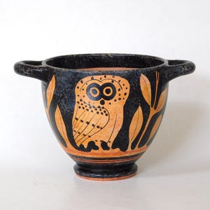 Könnte beinhalten: Eine schwarz-orangefarbene Keramikschale mit zwei Griffen. Die Schale zeigt ein stilisiertes Eulenmuster in Orange auf schwarzem Hintergrund. Die Eule ist von orangefarbenen blattartigen Formen umgeben. Die Schale hat einen abgerundeten Boden.