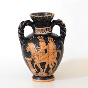 Può includere: Vaso in ceramica nera con una dettagliata illustrazione di figure e un carro trainato da cavalli in arancione e bianco. Il vaso ha manici attorcigliati e un bordo decorativo. Il design ricorda la ceramica greca antica.